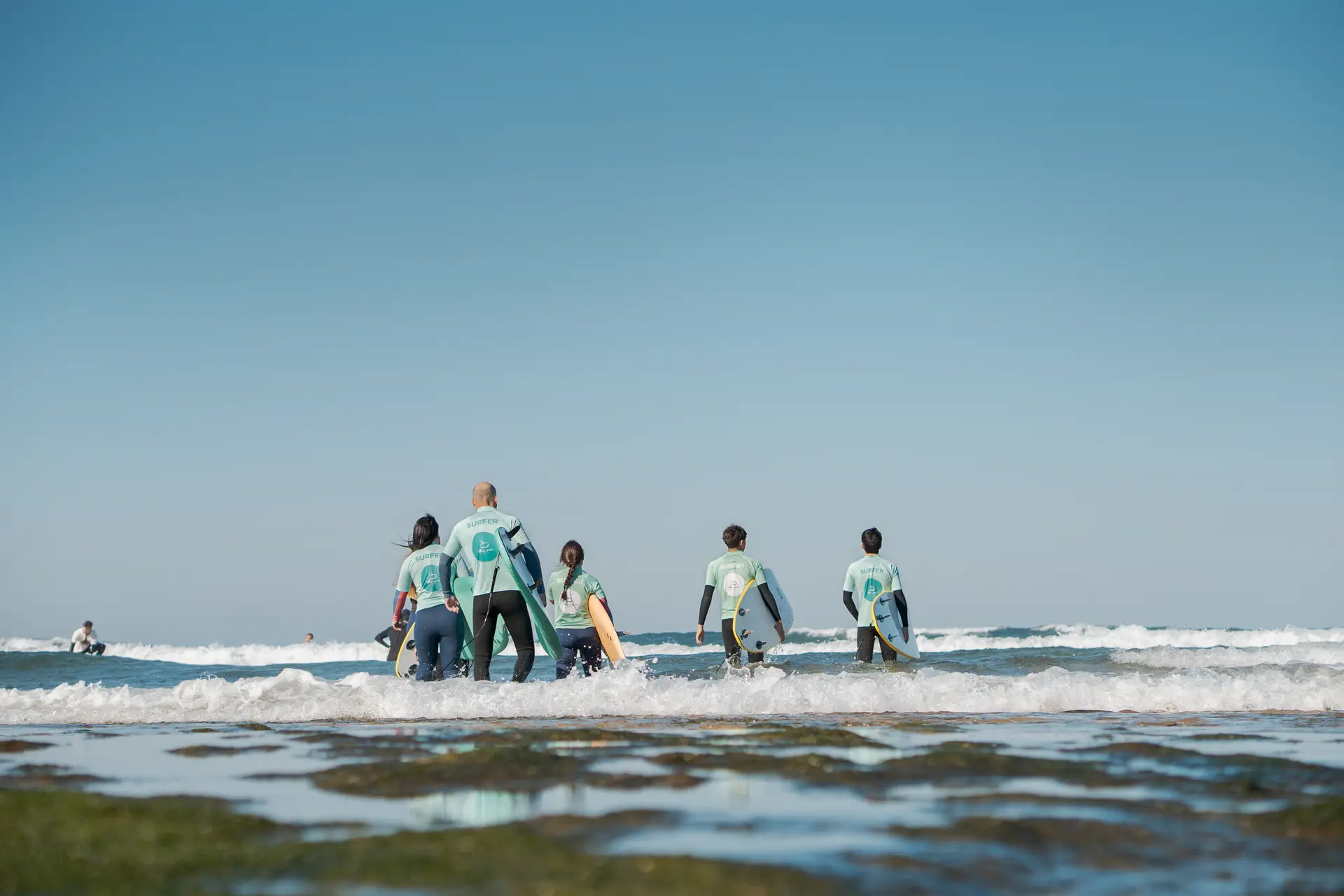 Alunos de surf a entrar no mar com as pranchas